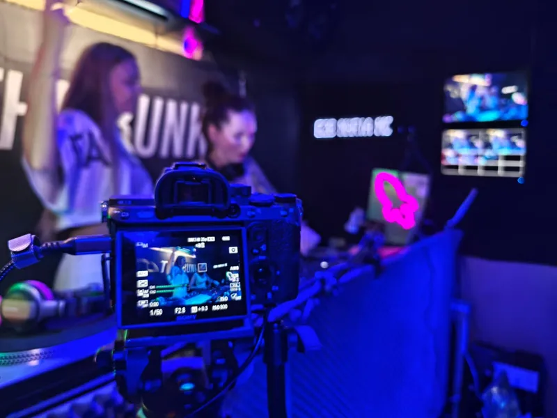 Pantalla de la cámara encuadrando a las DJs en la cabina, con los monitores multicámara al fondo