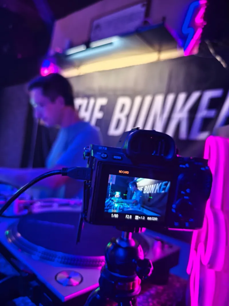 Cámara grabando a un DJ desde detrás de los platos, con luz de neón